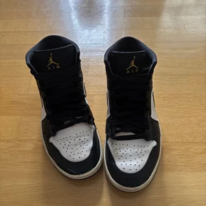 Nike Air Jordan 1 Mid svart/vit/guld - Snygga Nike Air Jordan 1 Mid sneakers i svart, vitt och guld. Klassisk siluett med svart snörning, perforerad tå och guldiga detaljer på swoosh och häl. Skorna har en robust svart yttersula och är tillverkade i skinn och syntet. Perfekta för dig som gillar streetstyle.