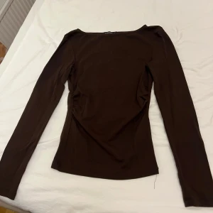 Brun långärmad topp från Stradivarius - Säljer en mörkbrun långärmad topp från Stradivarius i stretchig bomull. Toppen har en smickrande slim passform och lätt rynkade detaljer i sidan. Perfekt att styla med jeans eller kjol för en clean look.