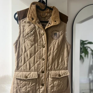 Beige väst från Lexington - Snygg beige quiltad väst från Lexington med bruna axelpartier och detaljer i manchester. Västen har tryckknappar, två stora fickor framtill och broderad logga på bröstet. 