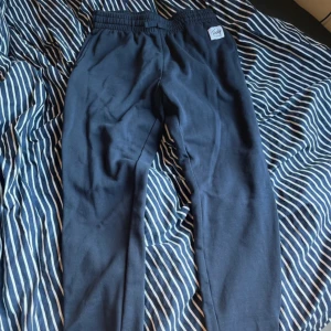 Mörkblå mjukisbyxor från Andy - Mörkblå mjukisbyxor från Andy med resår i midjan och mudd vid benslut. Byxorna har en enkel och clean look, sidofickor och en liten logga framtill. Perfekta för chill dagar eller när du vill vara bekväm.