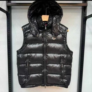 Svart dunväst från Moncler - Snygg svart dunväst från Moncler med glansig finish, avtagbar huva och två dragkedjefickor framtill. Västen har en hög krage och klassisk Moncler-logga på bröstet. Perfekt för lager-på-lager och streetstyle.