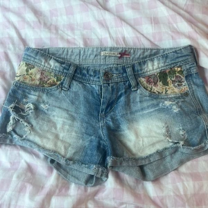 Blommiga jeansshorts från Stradivarius - Snygga ljusblå jeansshorts från Stradivarius med slitna detaljer och blommigt tyg på fickorna. Shortsen har rullad kant och en liten vinröd rosett vid etiketten. Perfekta för sommarens alla äventyr! Dragkedjan är trasig men det är lättfixat! Säljer för de är för små