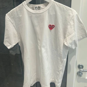 Vit t-shirt från Comme des Garçons - Säljer en stilren vit t-shirt från Comme des Garçons med det ikoniska röda hjärtat på bröstet. T-shirten är kortärmad och tillverkad i mjuk bomull. Perfekt för en avslappnad look. Storlek M men passar mer som S. Säljer för att den vart för liten för mig! 