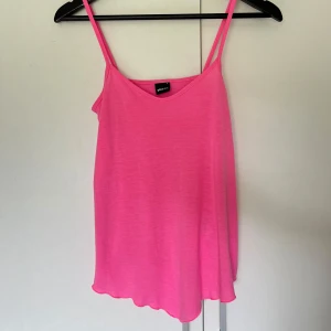 Rosa linnetopp från Gina Tricot - Supersöt rosa linnetopp från Gina Tricot med tunna axelband och avslappnad passform. Toppen har en enkel design med lätt v-ringning och är tillverkad i mjuk bomull, perfekt för varma dagar. Färgen är riktigt poppig och ger en härlig energi till din outfit.