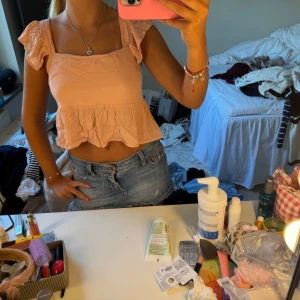 Ljusrosa volangtopp med knyt i ryggen - Supersöt ljusrosa topp med volangdetaljer och fyrkantig ringning. Toppen har korta volangärmar och en peplum-liknande nederdel. Baksidan har en öppen detalj med knytband. Perfekt för varma dagar och ger en romantisk vibe.