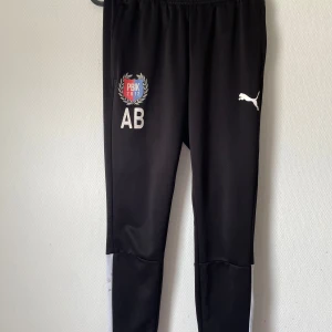 Svarta träningsbyxor från Puma - Svarta träningsbyxor från Puma med vitt tryck på ena benet och klubbmärke. Byxorna har smal passform, elastisk midja och vita detaljer längs benen. Perfekta för fotboll eller annan träning.