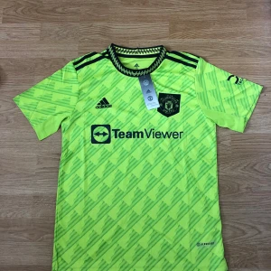 Manchester United Adidas matchtröja gul - Manchester Uniteds officiella matchtröja från Adidas i neon-gul med svart logga och detaljer. Tröjan har korta ärmar, diagonal mönstring och svarta axelband. Materialet är lätt och andas, perfekt för fotboll eller träning. TeamViewer sponsortryck på bröstet.