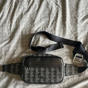 Svart magväska från Lacoste med mönster - Snygg svart magväska från Lacoste med geometriskt mönster och silverfärgade detaljer. Väskan har justerbart band med metallspänne, dubbla dragkedjor och präglad Lacoste-logga framtill. Perfekt för att bära det viktigaste med stil. Använd ett fåtal gånger, hör av er vid fler frågor. Pris kan diskuteras vid snabb affär.  