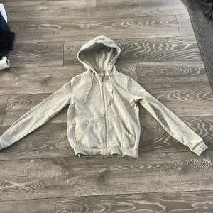 Grå hoodie med dragkedja från H&M - En basic ljusgrå hoodie från H&M med dragkedja framtill, huva med snörning och två fickor. Tillverkad i mjuk bomullsmix, perfekt för chill dagar. Långärmad och med en clean, enkel look som funkar till allt.
