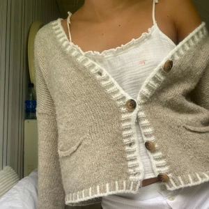 Beige stickad kofta från Stajl - Supermysig beige stickad kofta från Stajl med vita detaljer längs kanterna och stora träknappar. Koftan har två små fickor framtill och en loose passform. Perfekt att slänga över en topp för en chill look.