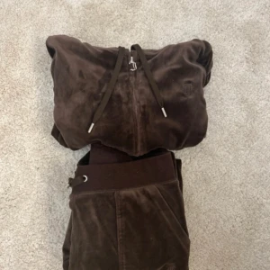 Brunt Juicy Couture sett - Mysigt brunt velour sett från Juicy Couture i xs på tröjan och s på byxan. Jackan har dragkedja framtill, huva och snörning. Perfekt för en chill och trendig look, klassisk Juicy-stil med mjukt material och ribbade muddar.