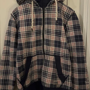 Rutig hoodiejacka från Jack & Jones - Snygg  hoodie från Jack & Jones i blått, vitt och rött. Jackan har dragkedja, huva med grova snören och två stora fickor framtill. Perfekt för dig som gillar en avslappnad och cool stil.