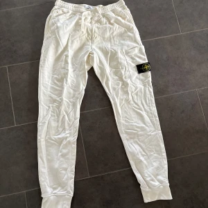 Vita mjukisbyxor från Stone Island - Snygga vita mjukisbyxor från Stone Island med klassisk patch på benet. Byxorna har dragsko i midjan, ribbade muddar och en praktisk cargoficka på sidan. Perfekta för en chill och avslappnad stil.     Fläckar vid höger ficka. 