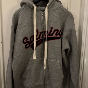 Grå hoodie med dragkedja från Salming - Snygg grå hoodie från Salming med vit snörning och dragkedja framtill. Stor broderad Salming-logga i svart och rött på bröstet. Mjuk insida och känguruficka. Perfekt för chill eller sportiga dagar.