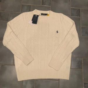 Vit stickad tröja från Polo Ralph Lauren - Säljer en stilren vit stickad tröja från Polo Ralph Lauren med klassiskt flätmönster. Tröjan har långa ärmar och en broderad logotyp på bröstet. Perfekt för en elegant och avslappnad look. Prislappen är dock borttagen