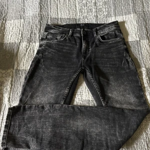 Svarta jeansbyxor från SMOG, 29/32 - Snygga svarta jeansbyxor från SMIG i tvättad look. Klassisk femficksmodell med dragkedja och knapp framtill. Jeansen har raka ben och tydliga slitningar för en cool, avslappnad stil. Perfekta för dig som gillar streetwear och vill ha en bekväm passform.