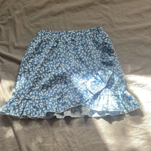 Blå blommig volangkjol från SHEIN - Supersöt blå kortkjol från SHEIN med vita småblommor och volangkant. Kjolen har omlott-look framtill och är gjord i lätt polyester, perfekt för sommarens alla dagar. Resår i midjan gör den bekväm och enkel att bära.