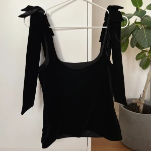 Svart sammetslinne med rosett från Zara - Supersnygg svart linnetopp från Zara i mjuk sammet. Breda axelband med stora rosetter som knyts på axlarna ger en unik och trendig look. Toppen har en rak, något croppad passform.