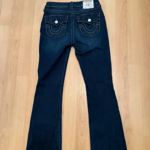 True Religion Becca Bootcut Jeans - Mörkblå jeans från True Religion, modell Becca Mid Rise Bootcut. Snygga kontrastsömmar och ikoniska fickdetaljer med knappar bak. Klassisk bootcut-passform och midja, tillverkade i mjukt jeansmaterial. 