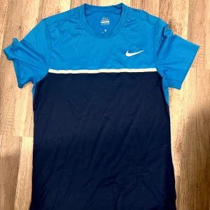 Nike blå Dri-Fit t-shirt S - Nike Dri-Fit t-shirt i sportig design med ljusblå överdel och mörkblå nederdel, avdelad med en vit linje över bröstet. Klassisk Nike-logga på bröstet. Tillverkad i lätt och snabbtorkande polyester, perfekt för träning eller tennis.