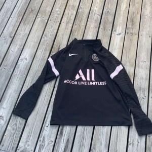 Svart PSG långärmad träningströja Nike - Svart långärmad fotbollströja från Nike och Paris Saint-Germain med rosa detaljer och klubbmärke på bröstet. Tröjan har halv dragkedja, tryckta loggor och texten 'Accor Live Limitless' samt 'Visit Rwanda' på ryggen. Tillverkad i funktionsmaterial som andas.VID SNABB KÖP KAN PRISET DISKUTERAS 