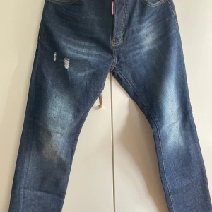 Dsquared2 blå jeans med patch - Snygga blå jeans från Dsquared2 med slitningar och coola detaljer. Baksidan har en stor gul och röd patch med siffran 2 och autograftryck. Jeansen har klassisk femficksdesign, orange sömmar och är tillverkade i bomullsdenim.