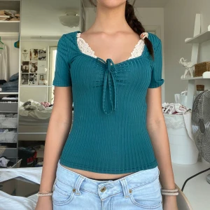 Turkos ribbad topp med spets och knyt - Supersöt turkos ribbad topp med kort ärm, v-ringning och vit spetskant upptill. Snygg knytning framtill som ger en feminin touch. Figurnära passform och stretchigt material, perfekt till jeans eller kjol.