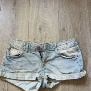 Ljusblå jeansshorts från H&M Divided - Snygga ljusblå jeansshorts från H&M Divided med upprullade benslut och slitna detaljer framtill. Klassisk femficksmodell med gylf och knapp, samt bälteshällor. Perfekta för varma dagar och en avslappnad look.
