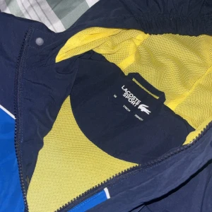Blå vindjacka från Lacoste Sport - Säljer en blå och marinblå vindjacka från Lacoste Sport med gul meshfoder. Jackan har dragkedja, hög krage och tydlig Lacoste-logga framtill. Perfekt för dig som gillar sportig stil och vill sticka ut med snygga färgblock. Jackan är i syntetmaterial och har normal passform. äkta köpt i butik. 1950kr riktigt bra skick ej använd nångång för att varan är för liten för mig
