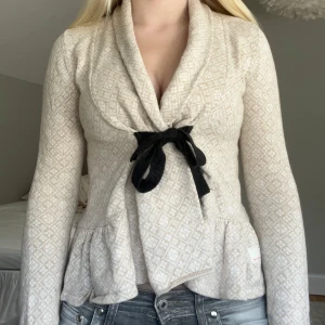 Beige kofta med svart rosett Odd Molly - Supersöt beige kofta från Odd Molly med diskret mönster, svart rosett framtill och broderad logga på ryggen. Koftan har lång ärm, omlottmodell och volangkant nertill. Perfekt för dig som gillar en feminin och chill stil.