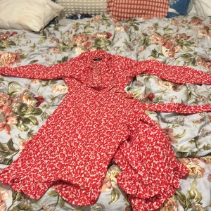 Röd blommig omlottklänning Amelie & Me - Supersöt röd omlottklänning från Amelie & Me med vitt blommönster, långa ärmar och volangdetaljer vid fållen. Perfekt för dig som gillar romantisk stil och vill sticka ut med färg. Klänningen har knytband i midjan och är lätt och luftig.