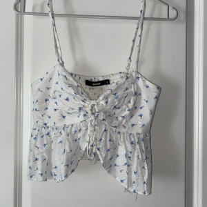 Vit blommig linnetopp från Bik Bok - Supersöt vit linnetopp med blått blommönster från Bik Bok. Toppen har smala axelband, knytning och rynkdetalj framtill samt volangig nederkant. 