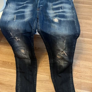 Dsquared2 slitna blå jeans - Riktigt snygga blå jeans från Dsquared2 med slitningar och trasiga detaljer på benen. Jeansen har en klassisk femficksmodell, coola slitningar både fram och bak samt Dsquared2-patch bak i midjan. Perfekt för dig som gillar en edgy streetstil.