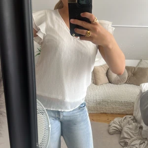 Vit blus med volangärmar - Säljer en söt vit blus med v-ringning och korta volangärmar. Blusen har en lätt textur i tyget och är luftig, perfekt för varmare dagar. Passar snyggt till både jeans och kjol för en fräsch look.