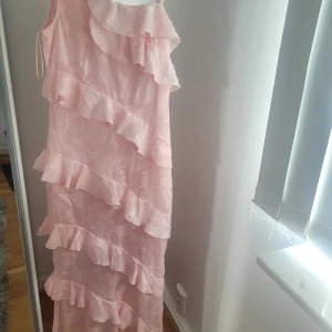 Ljusrosa långklänning med volanger - Superfin ljusrosa långklänning med volanger i flera lager och diskret blommönster. Klänningen har smala axelband och en romantisk vibe, perfekt för speciella tillfällen. Tunn och flowy känsla.