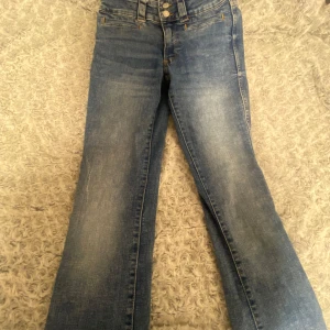 Blå bootcut jeans med dubbla knappar - Snygga blå jeans med bootcut passform och dubbla knappar i midjan. Jeansen har klassiska fickor bak med lock och knapp, samt detaljerade sömmar. Perfekta för dig som gillar en retro vibe och vill ha ett par jeans som sticker ut lite extra. Utsvängda. 