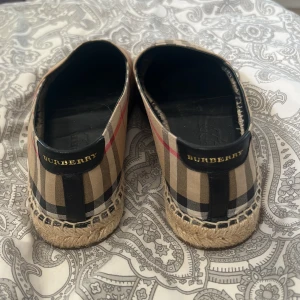 Burberry skor - Snygga espadrillos från Burberry med klassiskt beige, svart, rött och vitt rutmönster. Skorna har en platt sula och är Perfekta för en trendig sommarlook. De är använda max 3/4 gånger så helt nytt skick 💕