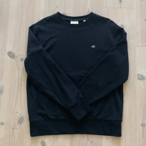 Svart sweatshirt från GANT - En stilren svart sweatshirt från GANT med rund halsringning och diskret logga på bröstet. Perfekt för en clean och enkel look.