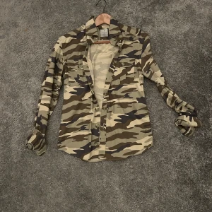 Camouflage skjorta med knappar - Snygg långärmad skjorta från la chemis i klassiskt camouflage-mönster med gröna, beige och bruna toner. Skjortan har två bröstfickor med lock och knappar framtill. Perfekt för dig som vill sticka ut med en cool militärstil. Storlek S eller 37