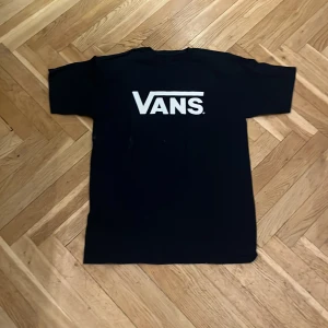 Svart Vans t-shirt med logga - Svart t-shirt från Vans med stor vit logga på bröstet. Klassisk passform med rund hals och korta ärmar. Perfekt för dig som gillar streetwear och enkel stil.