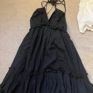 Svart långklänning från H&M Divided - Svart långklänning från H&M Divided med smala axelband som knyts i nacken och volangdetaljer. Klänningen har en luftig passform och är tillverkad i mjuk bomull, perfekt för varma dagar. Enkel men ändå stilren look. Aldrig använd.