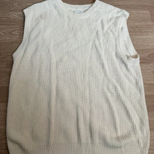 Stickad vit väst från East West - Säljer en stilren stickad väst från East West i en mjuk vit nyans. Västen har ribbad struktur, rund halsringning och är ärmlös. Perfekt att slänga över en skjorta eller t-shirt för en clean look.