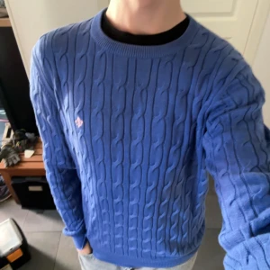 Kabelstickad Morris Tröja - Blå (Stl M) - Snygg blå kabelstickad tröja från Morris i storlek M. Tröjan är i gott skick men har en fläck på ena ärmen som kanske går bort i tvätt. Nypris ligger på ca 1600kr