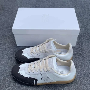 Maison Margiela sneakers med färgstänk - Unika sneakers från Maison Margiela i vitt skinn med svarta detaljer och coola färgstänk. Beige snörning och gummisula ger en retrovibe. Perfekta för dig som vill sticka ut med en konstnärlig touch på fötterna.