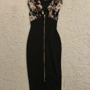Svart blommig klänning med dragkedja - Snygg svart långklänning från AX Paris med blommigt mönstrad överdel i ljusa och rosa toner. Djup v-ringning och ärmlös design. Klänningen har en guldig dragkedja framtill som ger en cool detalj. Figurnära passform som framhäver siluetten.