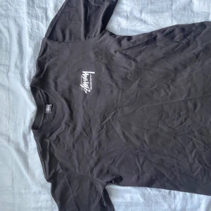 Svart Stüssy t-shirt med blommor - Svart t-shirt från Stüssy med vit logga på bröstet och stor print på ryggen med blommor i rosa och vitt samt texten 'Summer 19 No Bad Days Laguna Beach, CA'. Klassisk rund hals och korta ärmar. Skön bomullskvalitet och loose fit. Ikke autentisk 