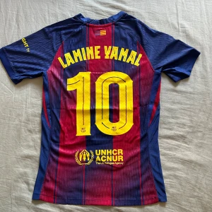 FC Barcelona Lamine Yamal #10 Nike tröja - FC Barcelona matchtröja med Lamine Yamal och nummer 10 på ryggen. Klassisk randig design i blått och rött med gula detaljer, klubbmärke och Nike-logga på bröstet. Tillverkad i lätt Dri-FIT-material, kortärmad och med sponsorer som Spotify och UNHCR tryckta på framsidan och ryggen.