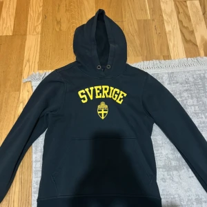 Svart Sverige-hoodie från Clique XS - Svart hoodie från Clique i storlek XS med gul text 'Sverige' och svensk vapensköld på bröstet. Klassisk känguruficka framtill och huva med snörningshål. Perfekt för dig som vill visa ditt stöd för Sverige på ett stilrent sätt.