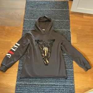 Mörkgrå Vlone hoodie med tryck - Säljer en grå hoodie från Vlone med stor V-tryck och motiv på framsidan, SVINT tryck på arm, samt texten 'Bless me father for I have sinned' i vitt och rött på ryggen. Hoodien har långa ärmar och huva, tillverkad i mjukt syntetmaterial. Perfekt för dig som gillar statement-plagg.