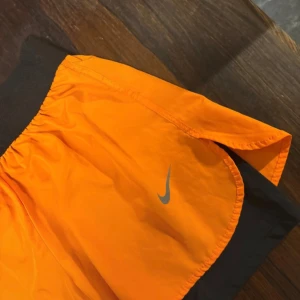 Orange träningsshorts från Nike - Snygga orange träningsshorts från Nike med svart elastisk midja och innerbyxa. Lätta och luftiga med klassisk Nike-logga i silver på framsidan. Perfekta för gymmet eller löprundan, med loose passform och sportig vibe.